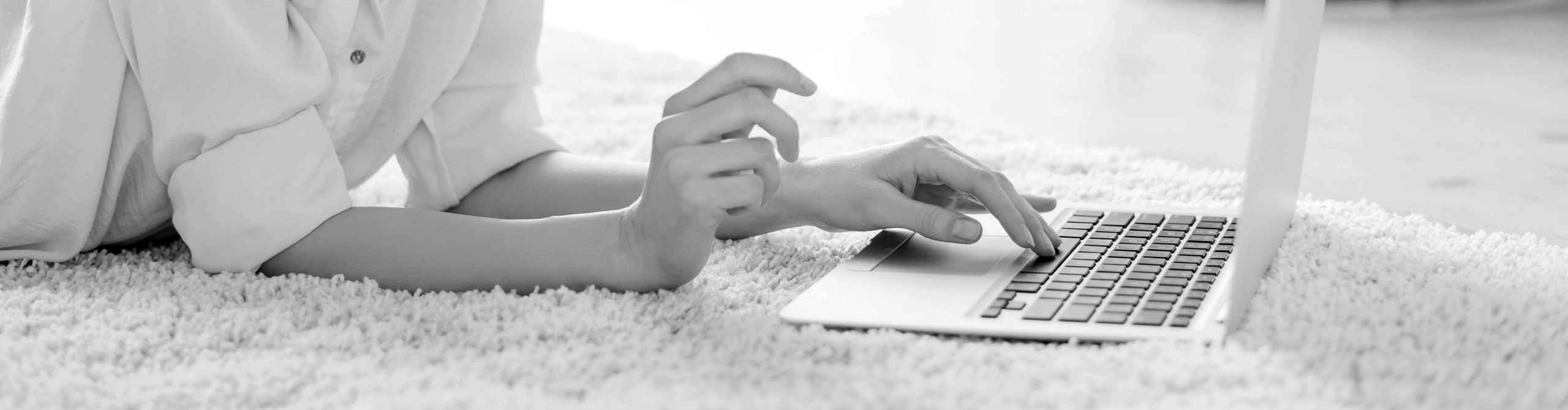 Person liegt auf einem flauschigen Teppich und tippt auf einem Laptop in einem hellen Raum. Header image